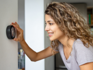 A smiling woman with curly hair adjusts the temperature on a smart thermostat in her home. This image illustrates the importance of comfort and control when choosing the right HVAC system for your home