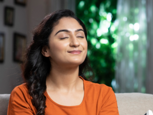 A relaxed woman in an orange shirt sits comfortably with her eyes closed, enjoying the fresh air inside her home. This image represents the importance of Indoor Air Quality for creating a healthy and comfortable living environment.